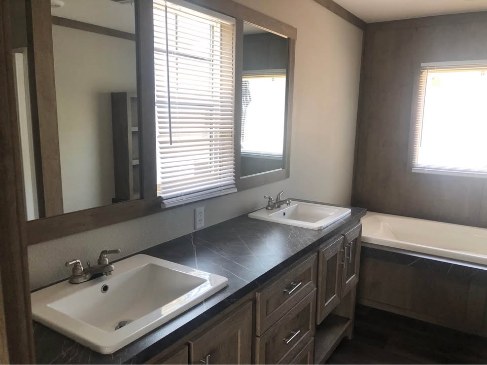 Modern bathroom with dark marble countertop featuring dual sinks and mirrors. A soaking tub is in the corner, with natural light streaming through blinds.