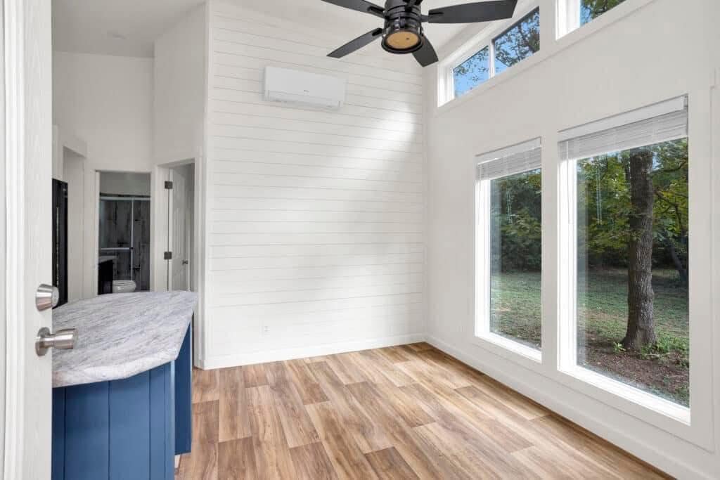 Bright room with white shiplap walls, large windows, and a wood floor. A ceiling fan hangs above, and a blue countertop is visible on the left.