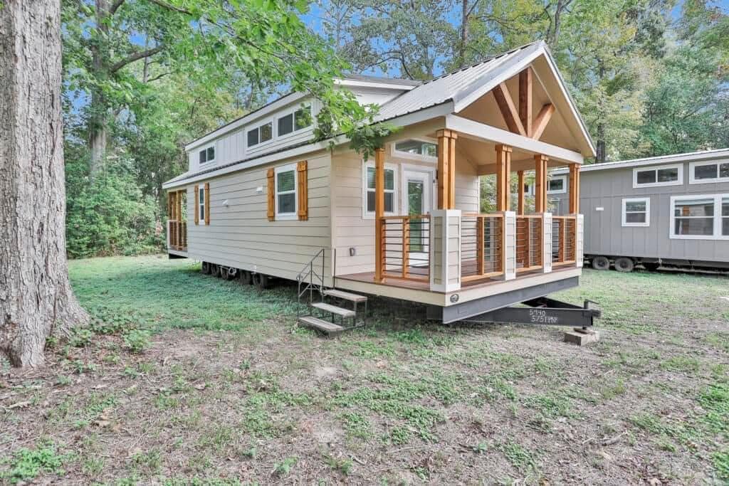 A charming tiny house with a beige exterior and wooden accents stands in a lush, wooded area. It features a small front porch and several windows, evoking a cozy and serene vibe.