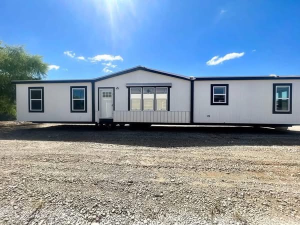 Single-story white manufactured home with a pitched roof on a gravel lot, under a clear blue sky. Bright sunlight casts shadows, creating a serene atmosphere.