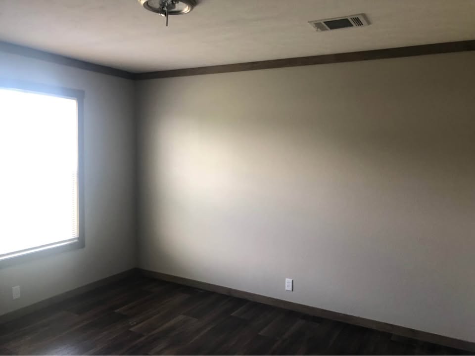 An empty room with a neutral beige wall, dark wood flooring, and a large window on the left. Soft light enters, creating a calm and minimalist atmosphere.