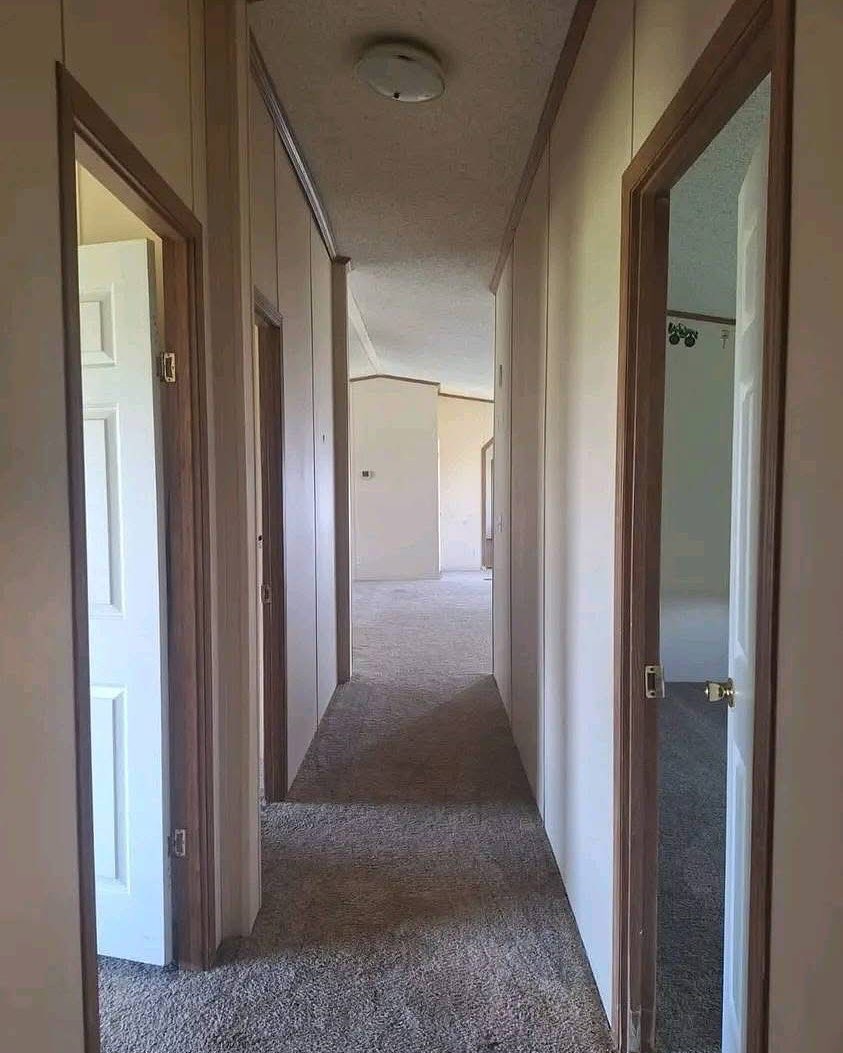 A narrow, carpeted hallway with beige walls and several open wooden doors leads to a softly lit room in the distance. The atmosphere is calm and empty.