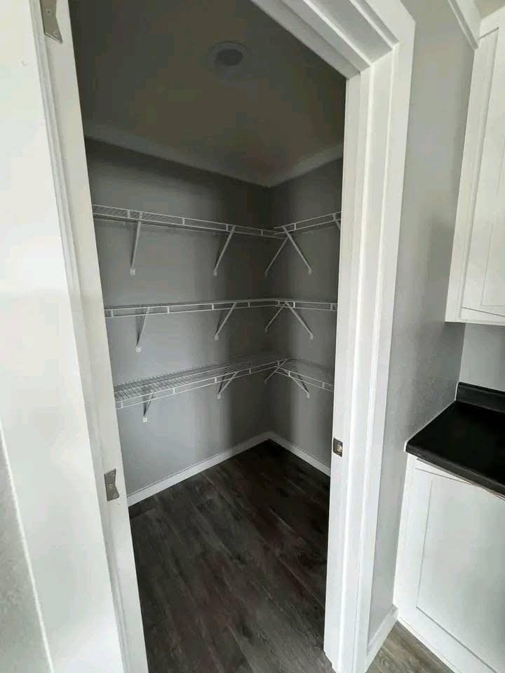 Empty walk-in pantry with light gray walls, dark wood flooring, and white wire shelving on three walls. Adjacent white cabinet with black countertop.
