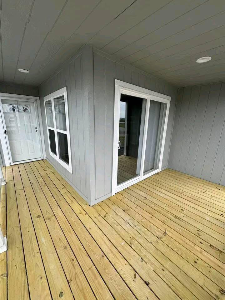Wooden deck with light gray siding on walls, featuring a glass sliding door and white-framed windows. Bright, clean, and modern outdoor space.