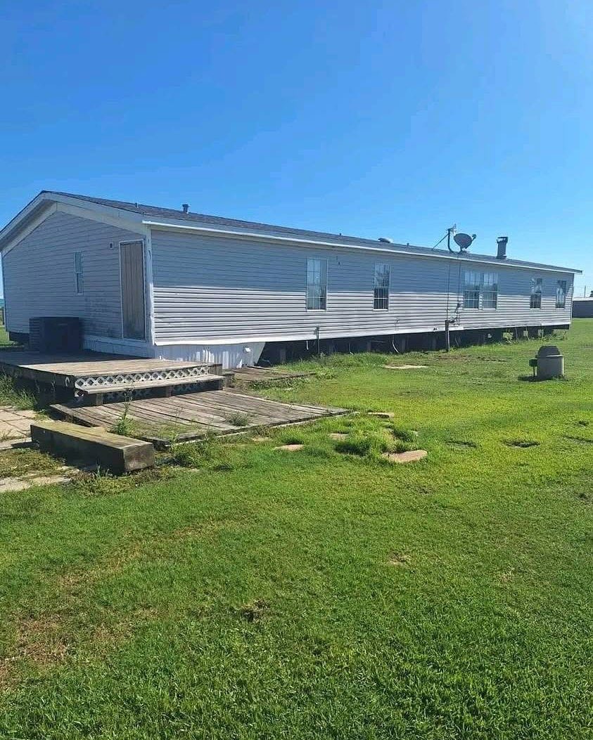 A long, white mobile home sits on a bright green lawn under a clear blue sky. A small wooden deck leads to the door, creating a serene, sunny atmosphere.