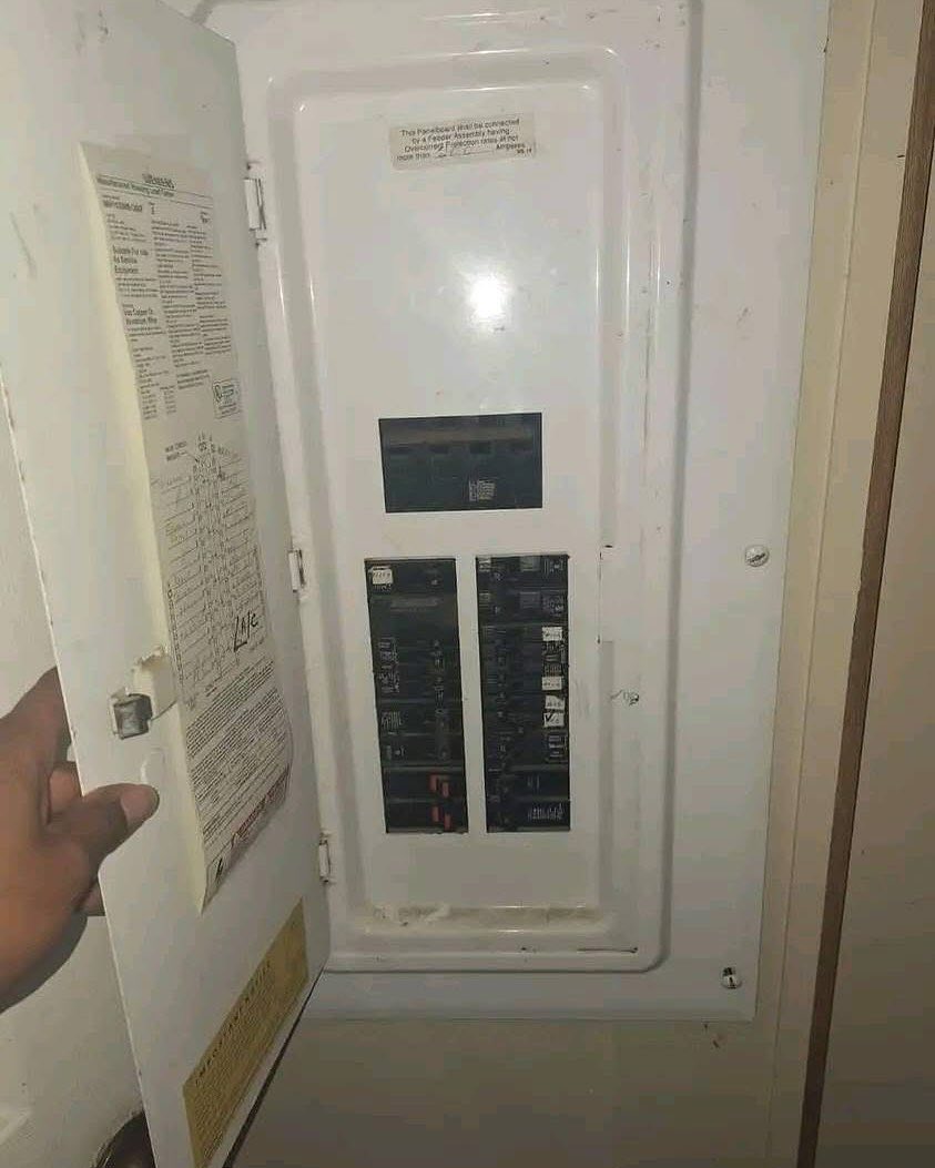 Open electrical panel box with a hand holding the door. Inside are rows of switches and labels. The panel is white and slightly worn.