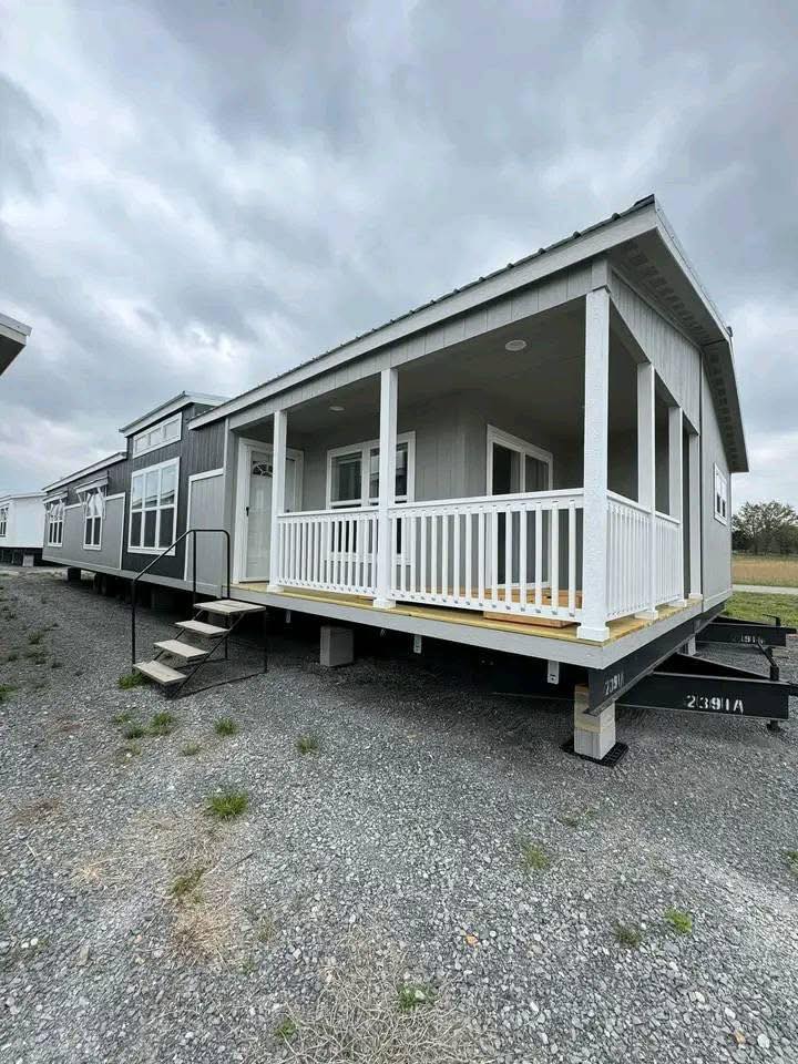Modern gray mobile home with a white porch railing under a cloudy sky. The structure is elevated with a small staircase leading to the porch.