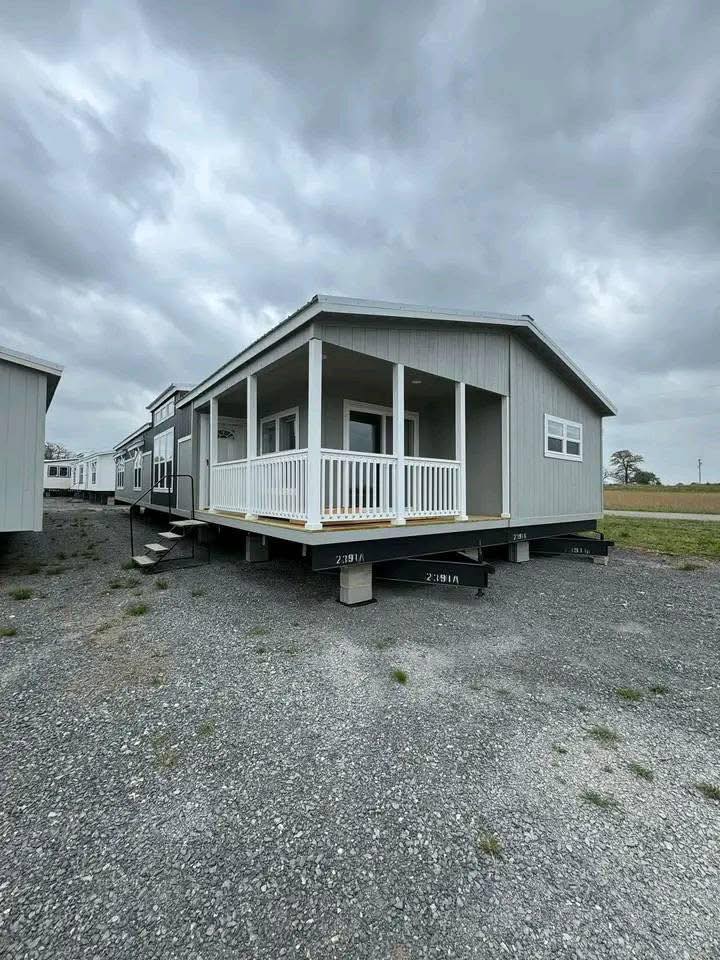 A gray manufactured home with a white porch sits on a gravel lot under a cloudy sky. Nearby are other similar homes, suggesting a sales or display area.