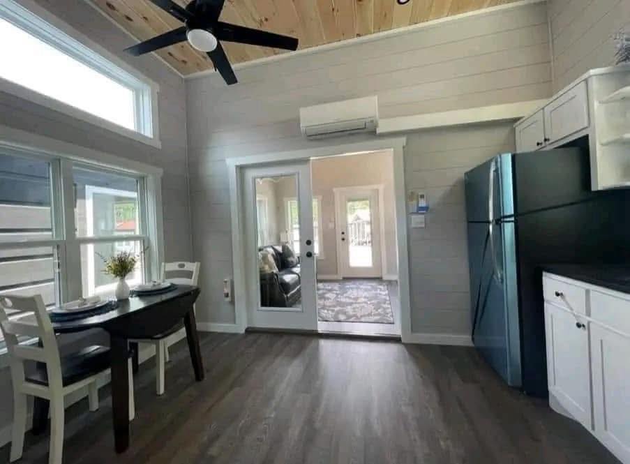Compact kitchen and dining area with light wood flooring, a dark table with white chairs, black fridge, and white cabinets. Glass doors lead to a living space.