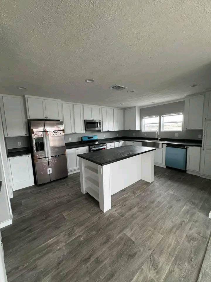 Modern kitchen with white cabinets, dark countertops, and stainless steel appliances. Features a central island and wooden flooring, creating a sleek, spacious feel.