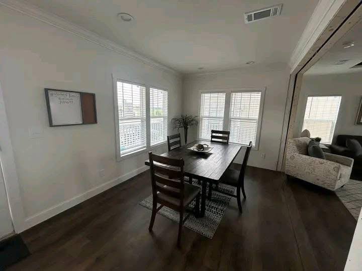 Dining room with dark wooden table and chairs, illuminated by natural light from large windows with blinds. A cozy, neutral-toned living area is seen through an open doorway, creating an inviting and warm atmosphere.