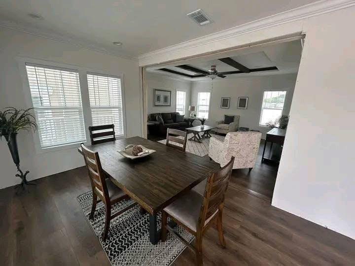 Spacious dining room with a dark wood table and four chairs on a patterned rug. Sunlit living area visible in the background, creating an open, airy feel.