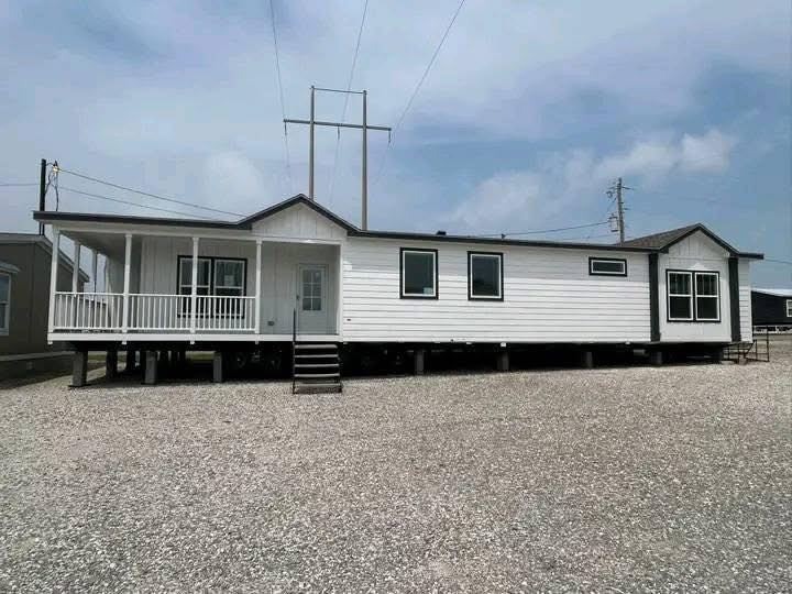 A white, elevated modular home with a front porch and multiple windows stands on a gravel lot under a partly cloudy sky, conveying a clean, serene atmosphere.