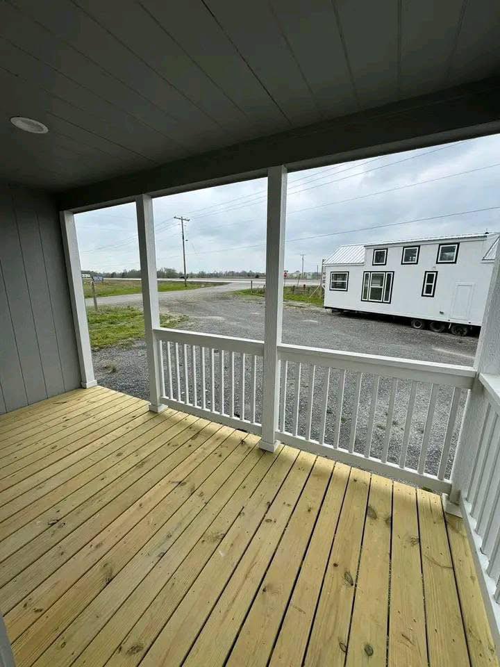 Covered wooden porch with white railings overlooks a gravel lot and a white tiny house. Overcast sky suggests a calm, subdued atmosphere.