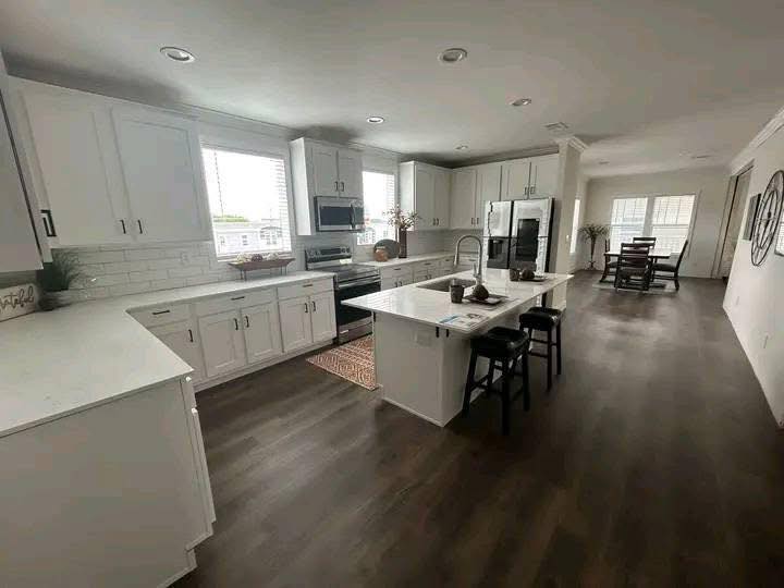 Modern kitchen with white cabinets, stainless steel appliances, and a central island with barstools. Warm wood floors add a cozy feel. Dining area visible in the background.