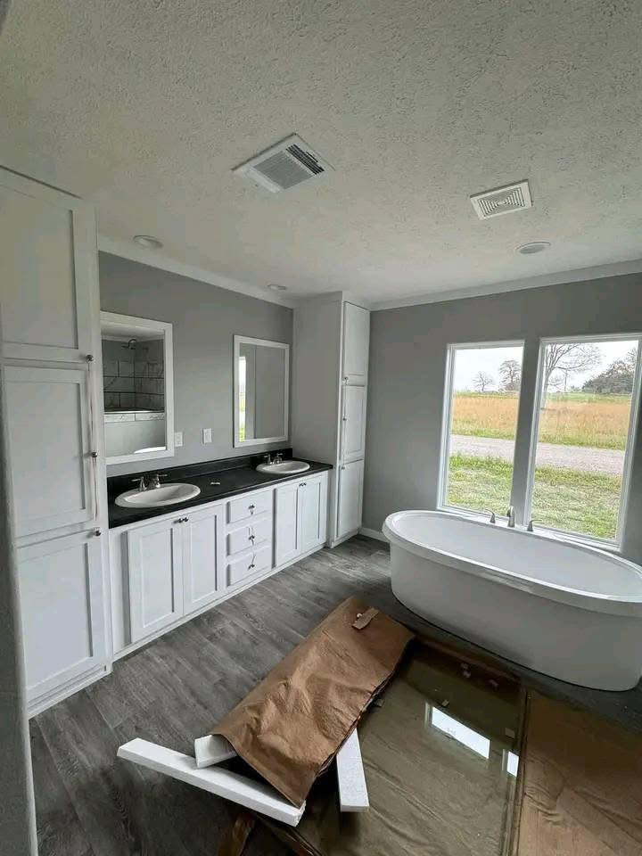 Modern bathroom with dual sinks on a black countertop, white cabinets, a freestanding tub by large windows, and wooden floors. A relaxed, serene tone.