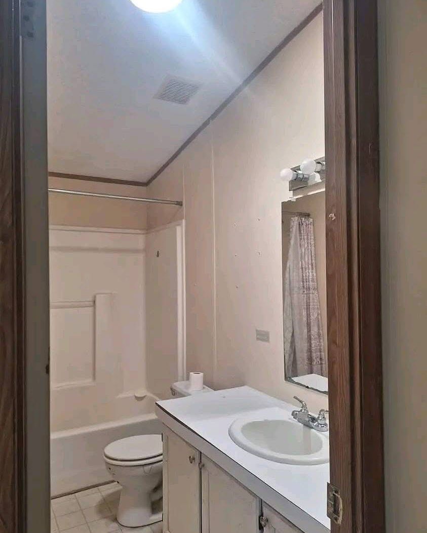 Small bathroom with beige walls, featuring a white bathtub with a shower curtain, a toilet, and a white vanity with a sink and mirror under bright light.