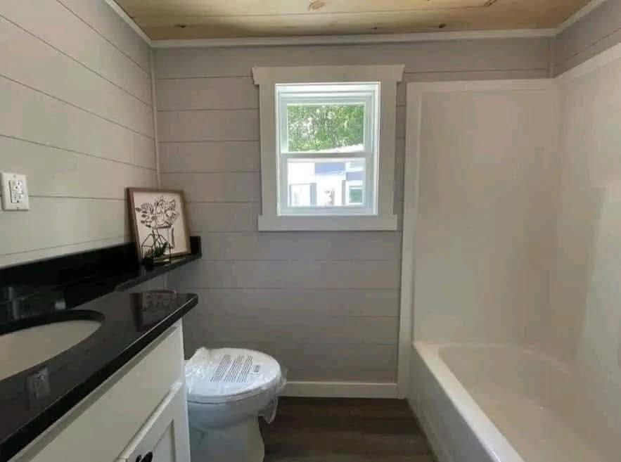 A small bathroom featuring a black countertop with a sink, a toilet, and a bathtub. Gray walls, white trim, and a window create a bright, fresh look.