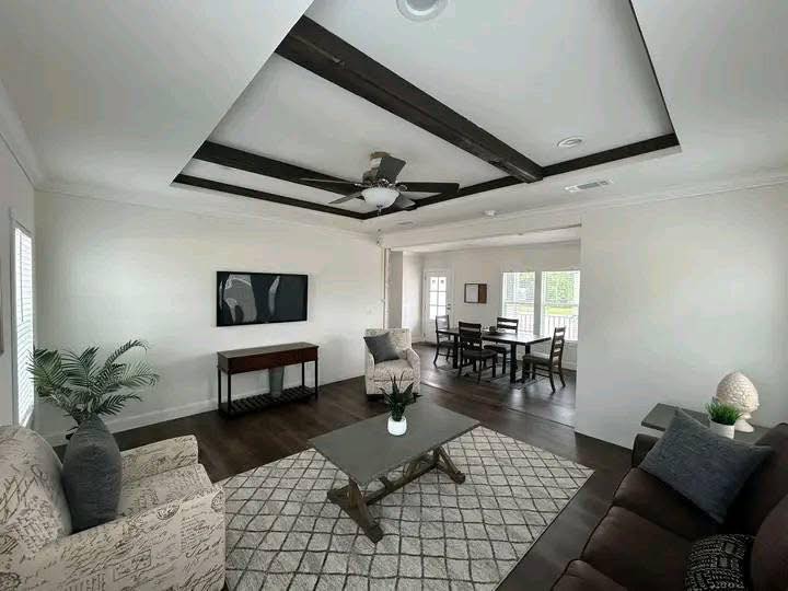 A modern living room with a geometric rug, gray armchairs, a brown sofa, a central coffee table, and an overhead fan. Dining area visible in the background.