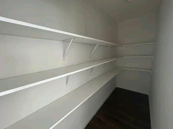Empty pantry with white shelves lining three walls, creating a clean and organized appearance. The room has a minimalist and spacious tone.