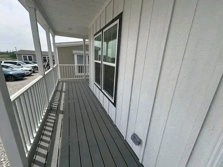 A long, wooden porch with gray floorboards and white railings extends alongside a white paneled house. Several parked cars are visible in the background.