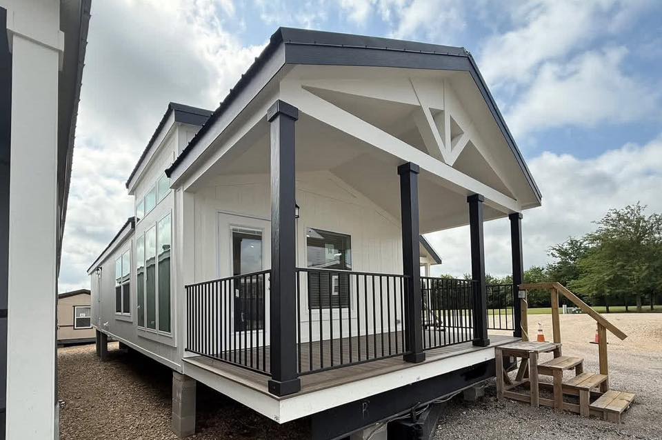 A modern tiny house with a white exterior, black trim, and a small porch with a railing. It sits on a gravel lot under a partly cloudy sky.