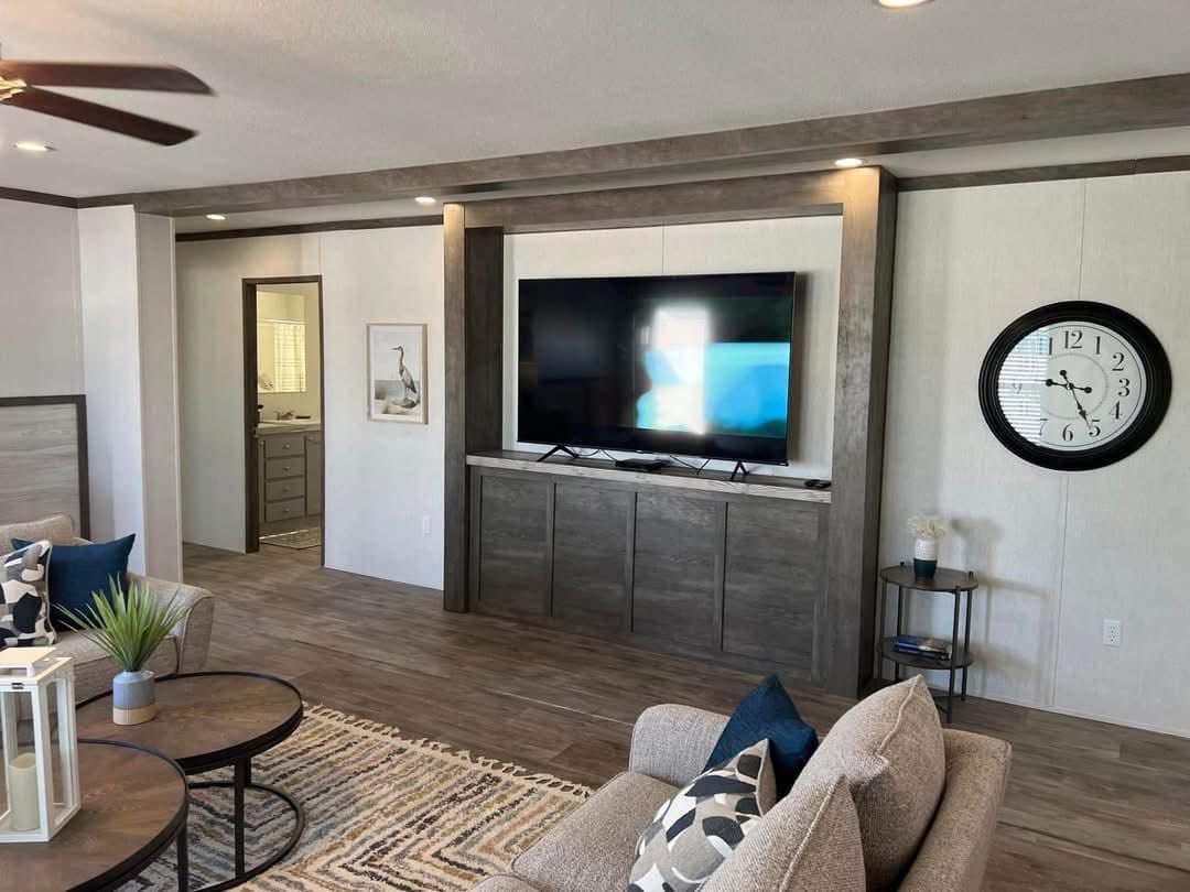 Spacious living room with a large TV on a wooden media wall, a round black-framed clock, comfy beige sofas with patterned cushions, and modern decor.