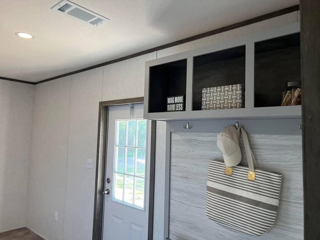A modern room corner with a white door, overhead shelf, and gray walls. A striped tote bag and a cap hang on a hook. The shelf holds baskets and a sign saying, "Wag more, bark less." The space is bright and minimalistic.