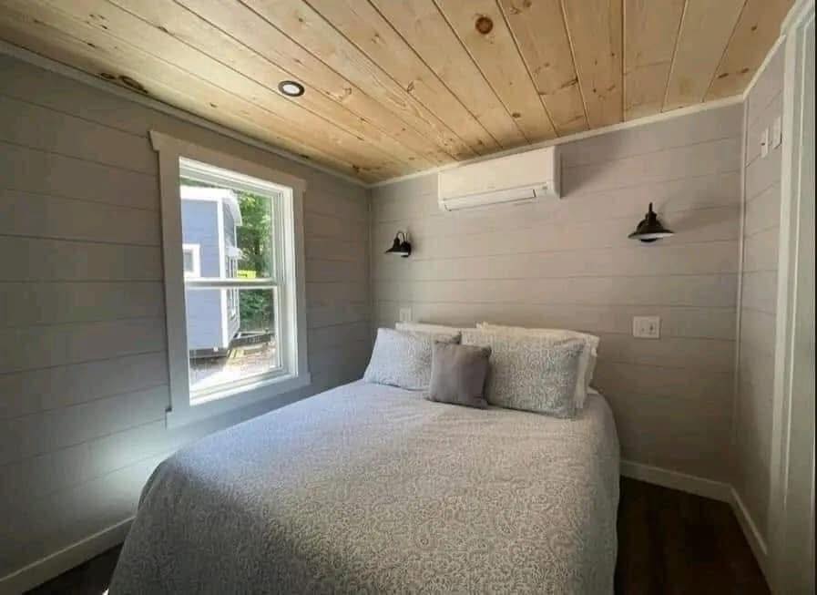 Compact bedroom with light gray walls and a wood-paneled ceiling. Features a double bed with patterned bedding, two pillows, wall sconces, and a window.