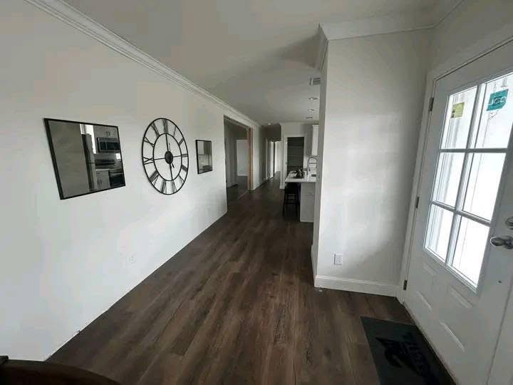 A bright hallway with hardwood floors leads to a kitchen. A large decorative clock and mirrors adorn the left wall, while a door with glass panels is on the right.