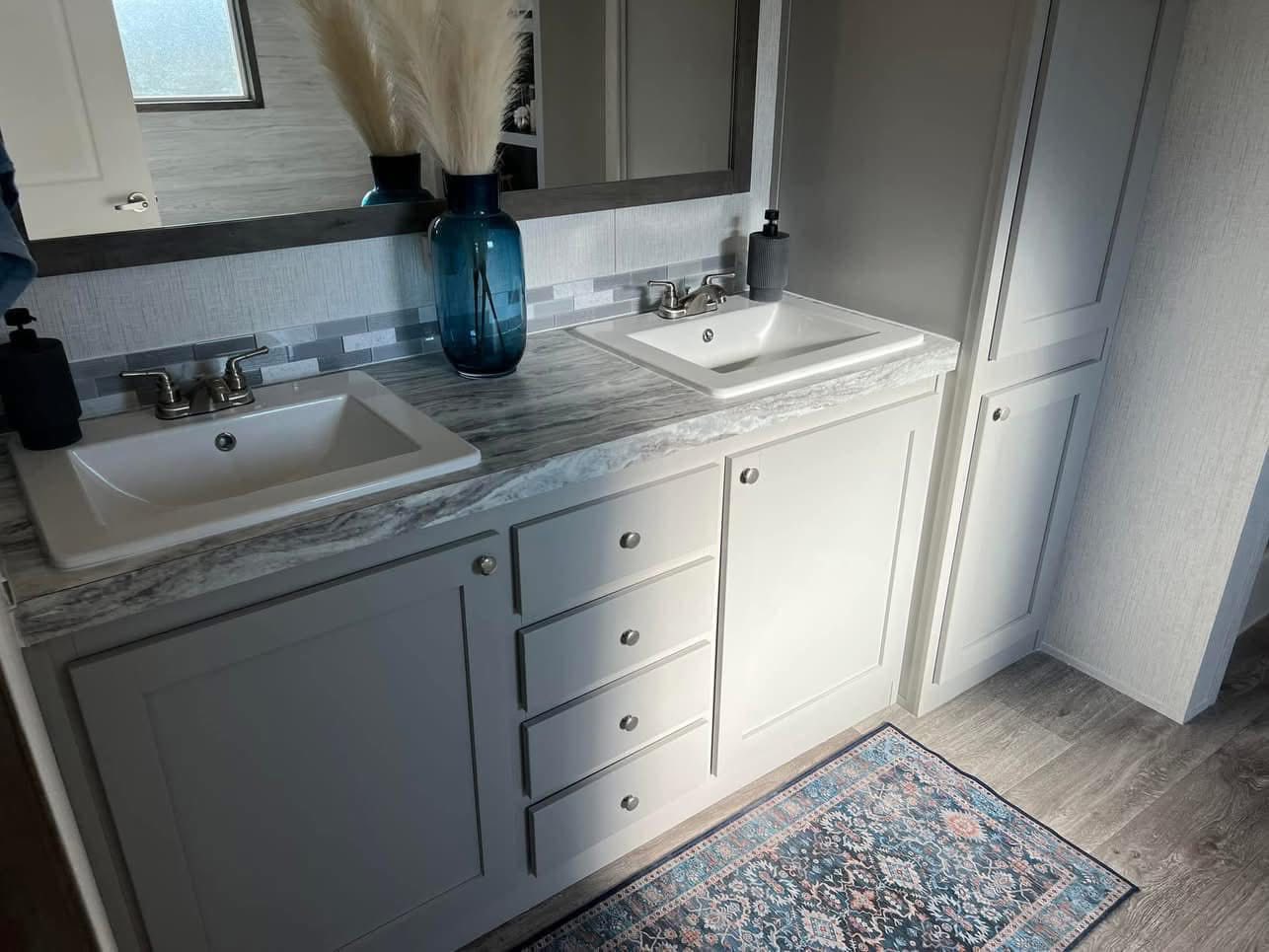 Bright bathroom with dual sinks on a gray marble countertop, white cabinetry, a large mirror, blue vase with pampas grass, and a patterned rug.