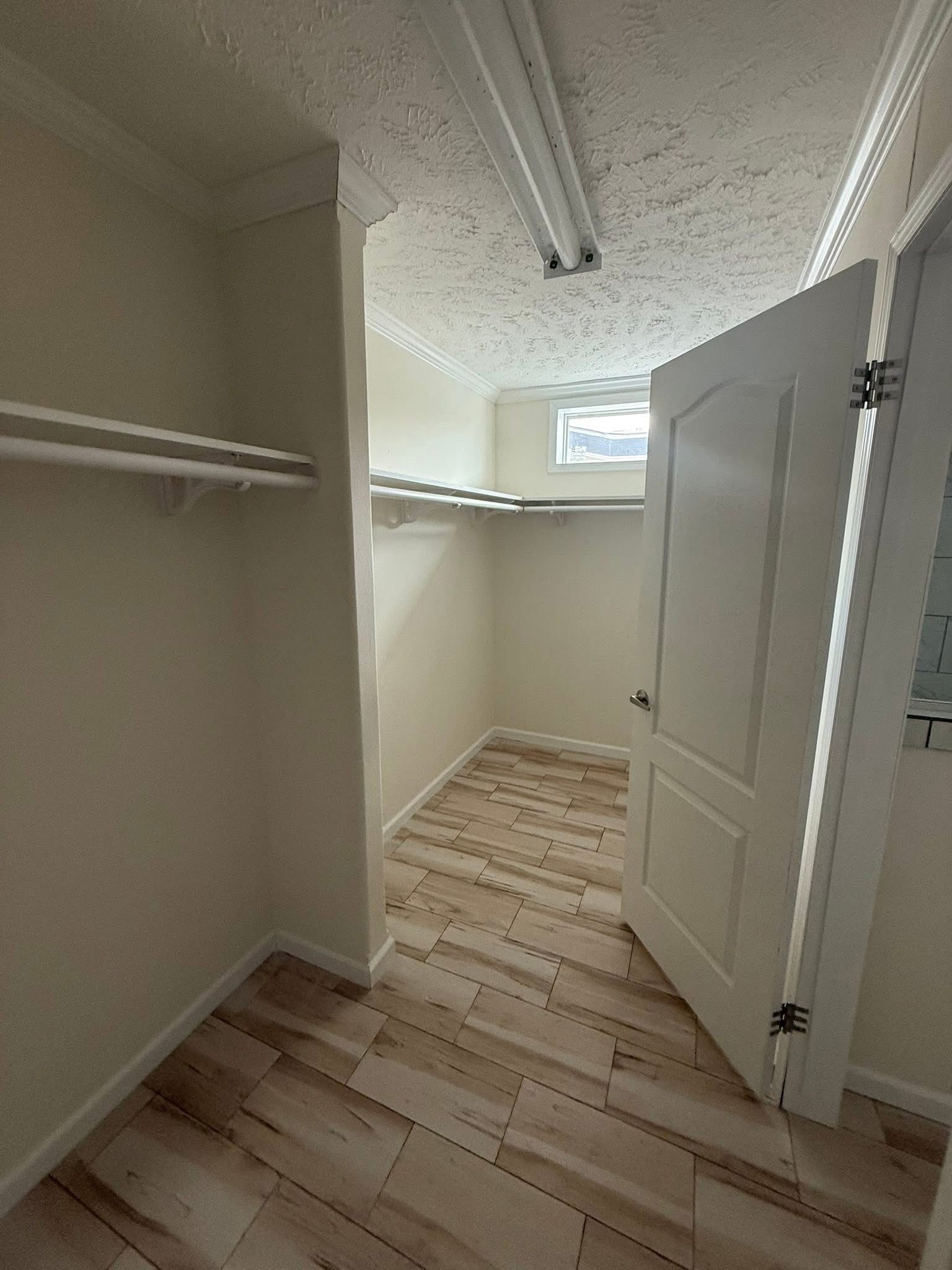Narrow walk-in closet with wooden tile floor, open door, fluorescent lights, and shelves on both sides. A small window brings in natural light.