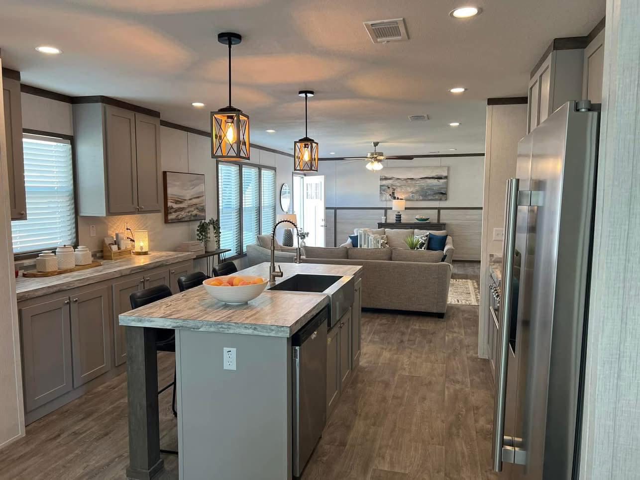 Modern kitchen and living room with gray cabinets, wood flooring, and neutral tones. Island with fruit bowl, pendant lights, and cozy seating area.