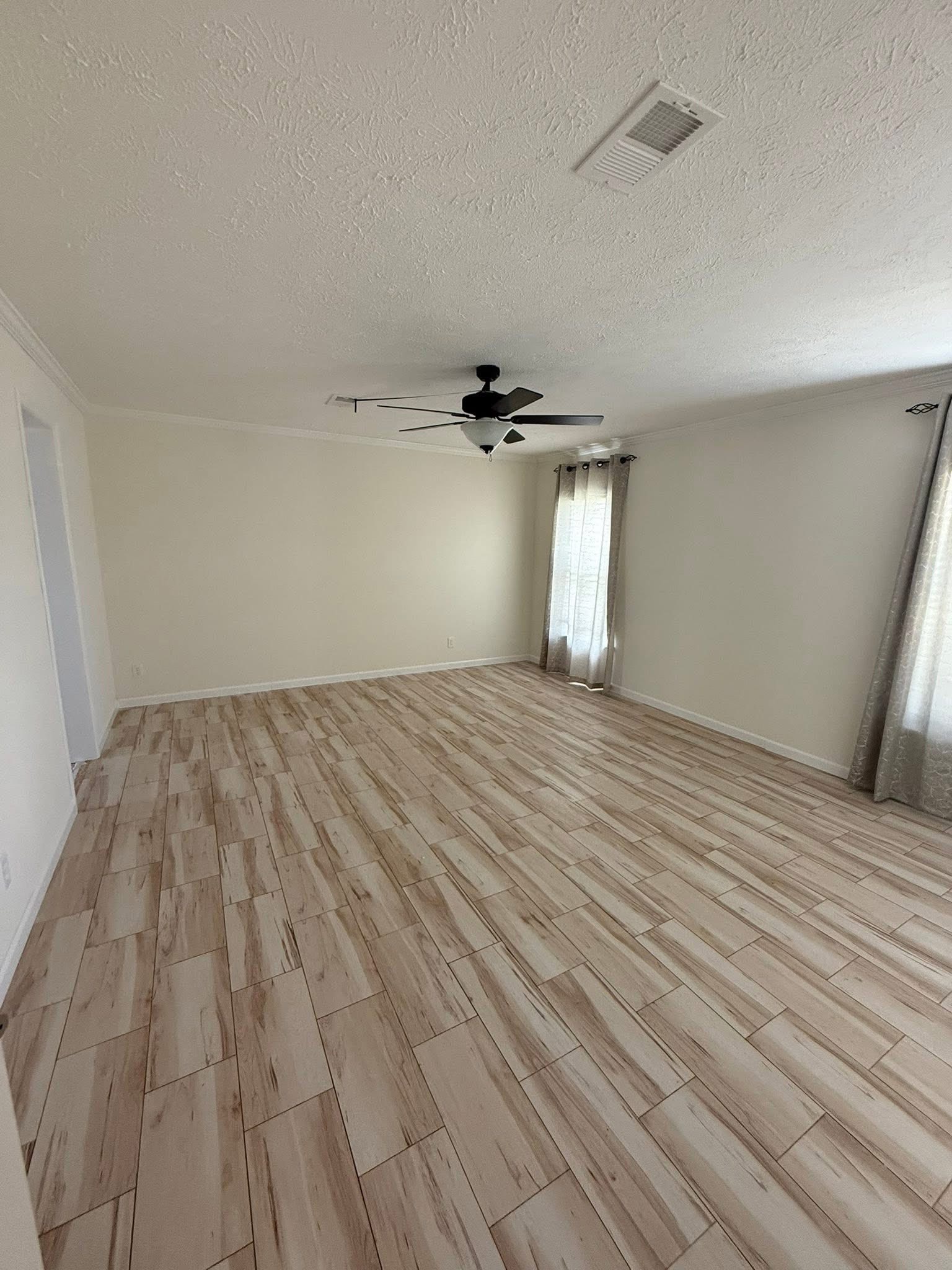A spacious, empty room with light wood flooring and cream-colored walls. A ceiling fan hangs from the white ceiling, and sheer curtains cover the window, adding a calm, airy feel.