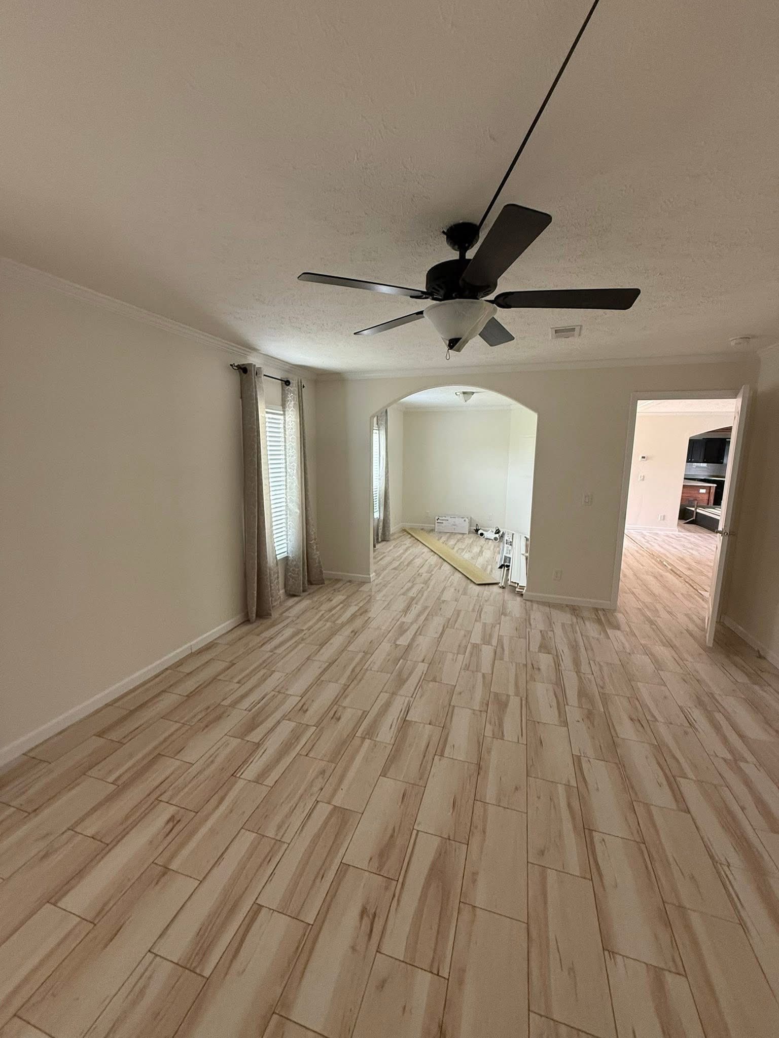 Spacious room with light wood-look tile flooring and neutral walls. A ceiling fan is centered above. Large windows with curtains and adjacent archway. Clean and modern ambiance.