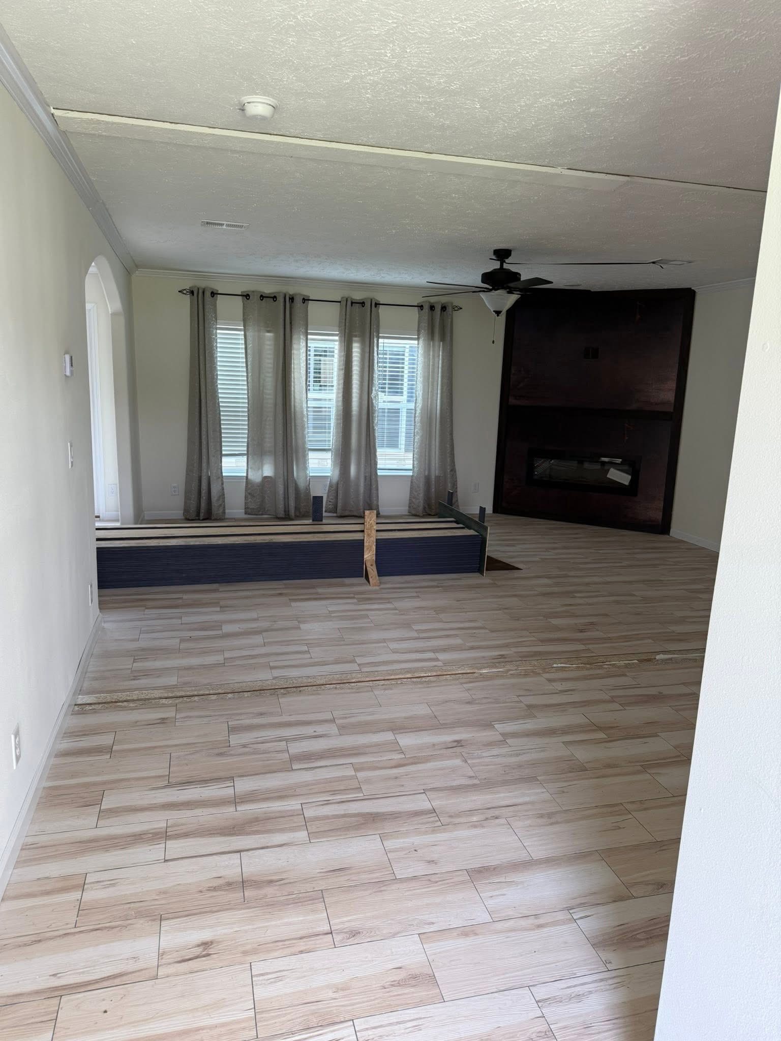 Spacious, empty living room with light wood floors, large windows with gray curtains, a ceiling fan, and built-in fireplace. Calm, minimalist ambiance.