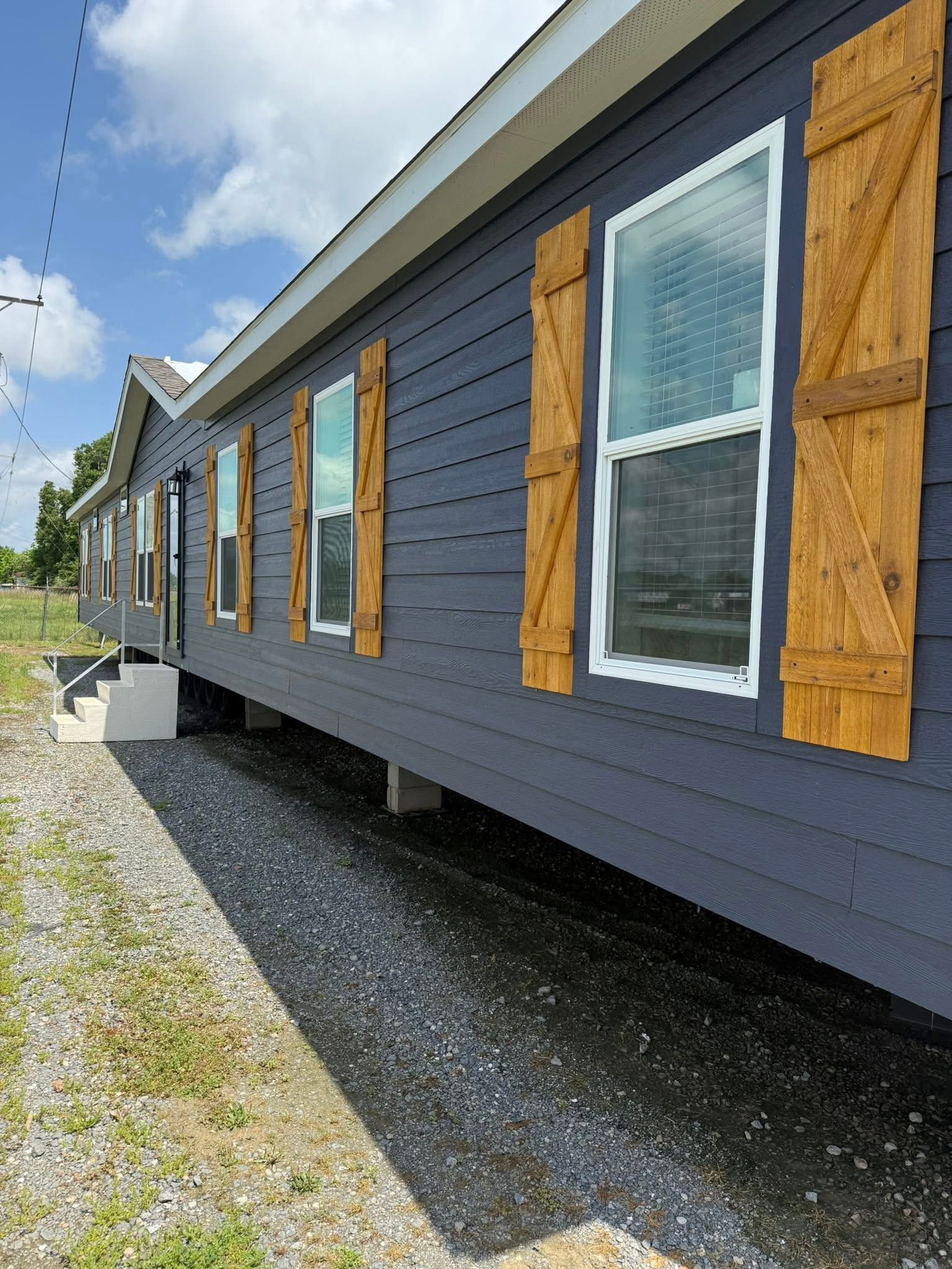 A dark blue house with wooden shutters, large windows, and a sloping roof under a partly cloudy sky. The gravel pathway leads to small stairs.