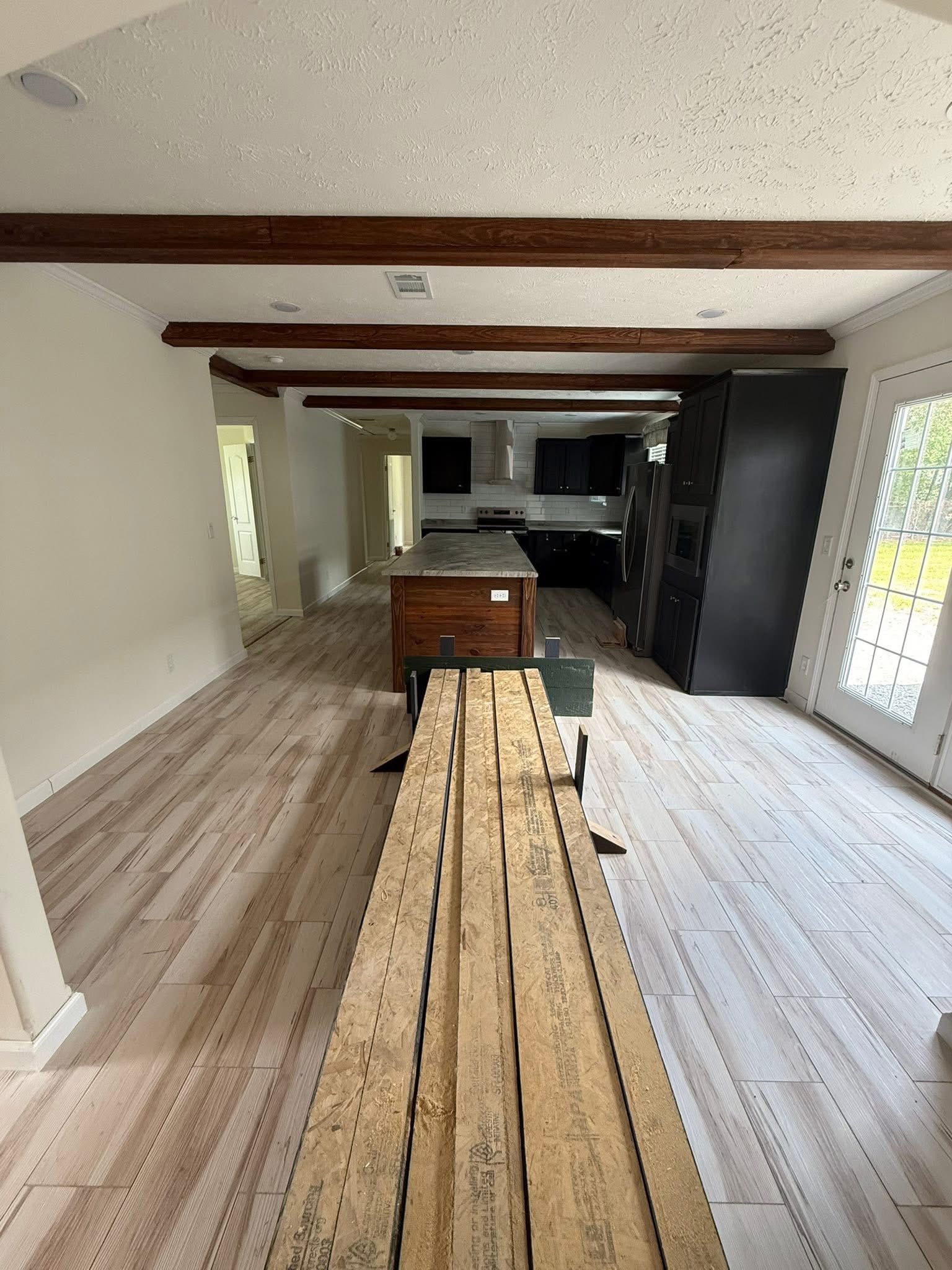 A spacious, light-filled kitchen under renovation, featuring exposed wooden ceiling beams, a long wooden work table, and modern black cabinets.