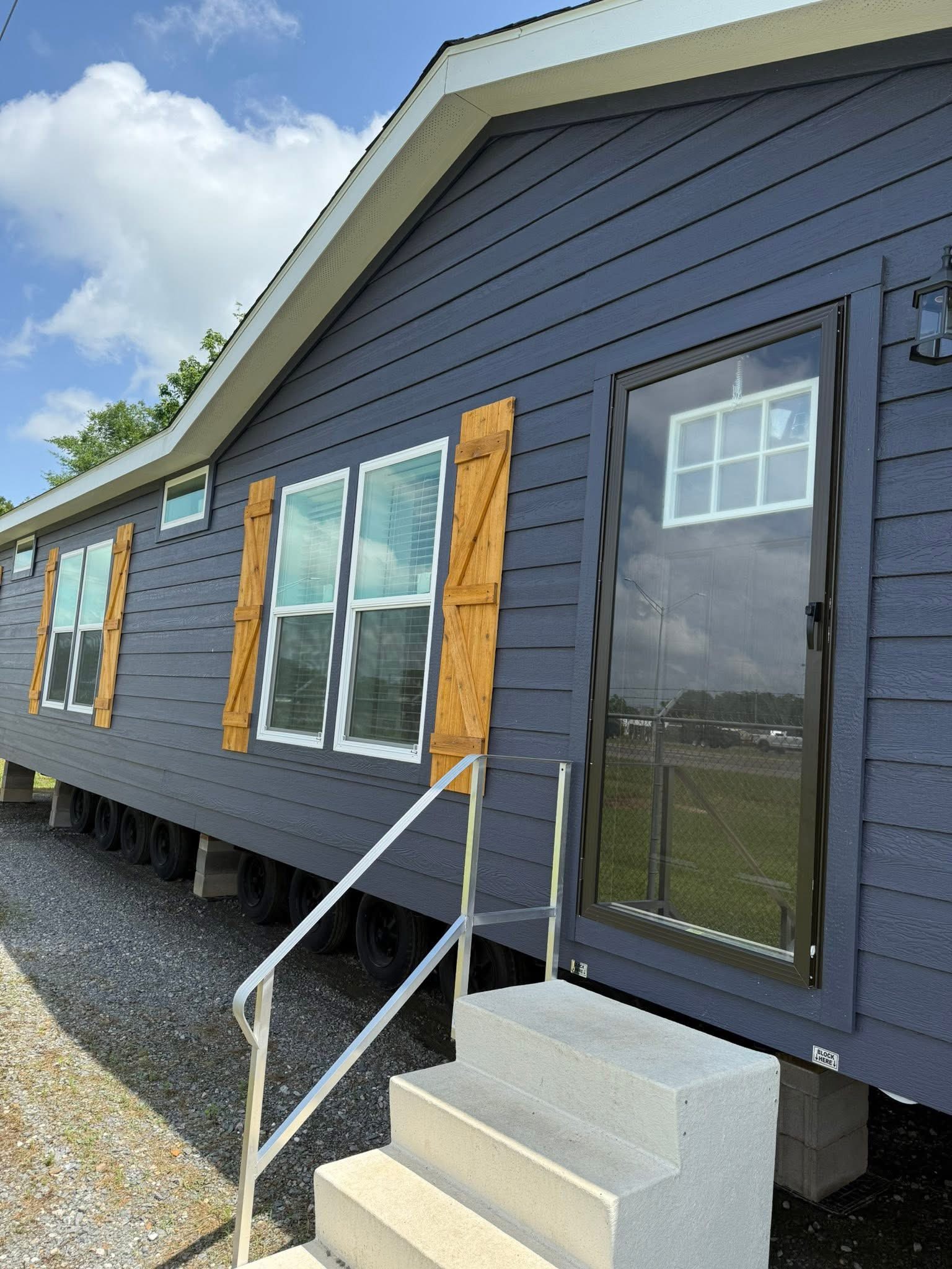 A modern blue mobile home with wooden shutters and large windows. A glass door leads to concrete steps with a metal railing. The sky is clear and sunny.