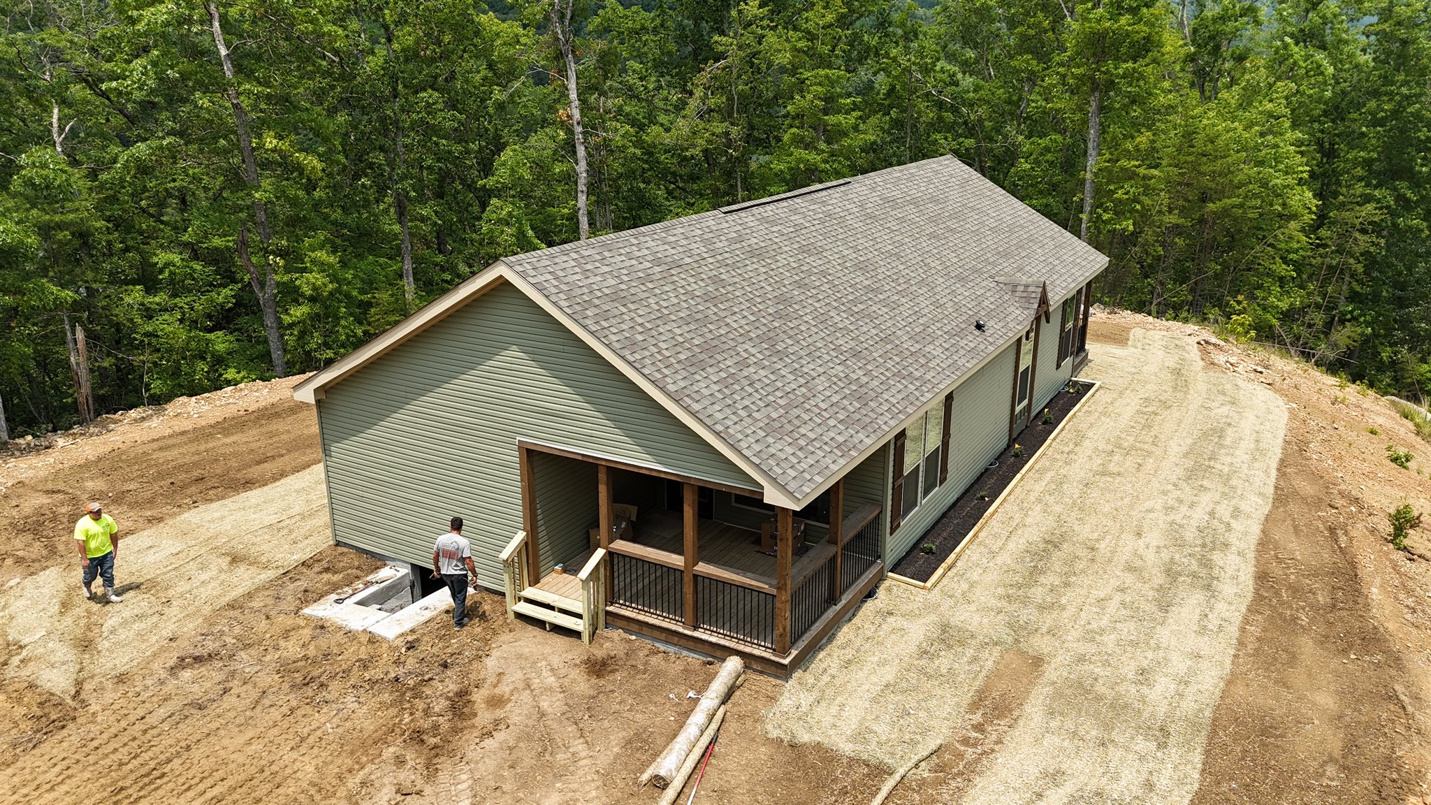 Aerial view of a single-story house with a gray roof surrounded by trees. Two people stand outside on a newly landscaped yard, conveying a sense of completion.