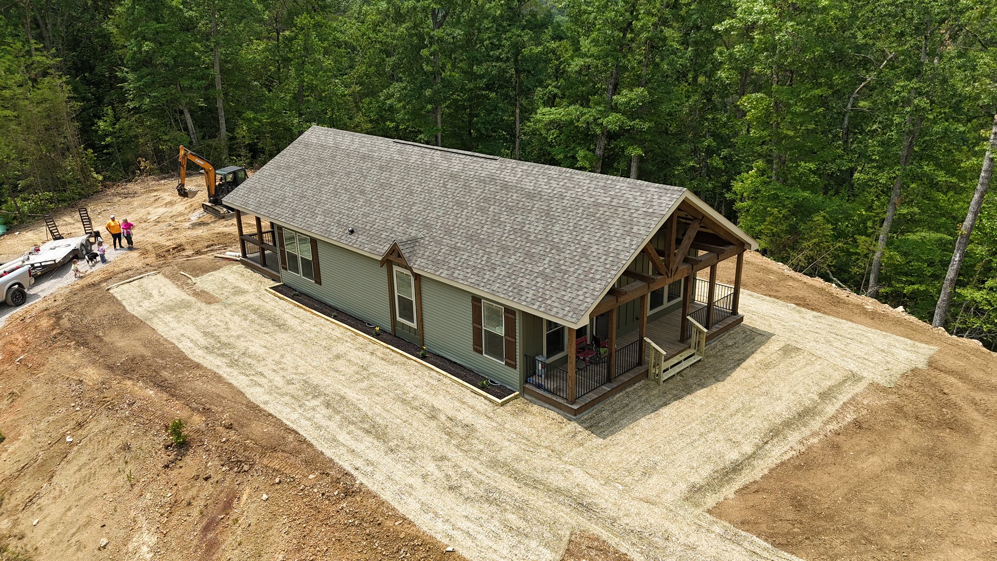 Aerial view of a new green house with a grey roof surrounded by trees. Workers and machinery, including a digger, are present on the dirt terrain.