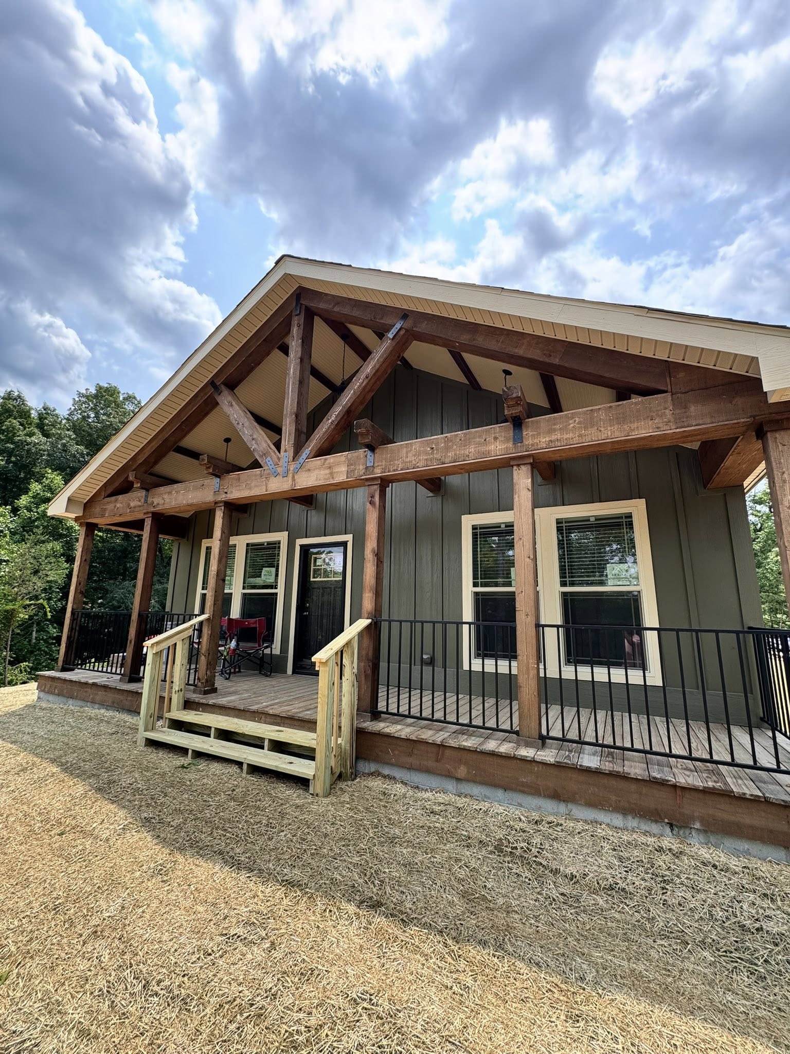 Rustic wooden cabin with a gabled roof and large porch, set against a cloudy sky. The peaceful setting is surrounded by trees and dry grass.