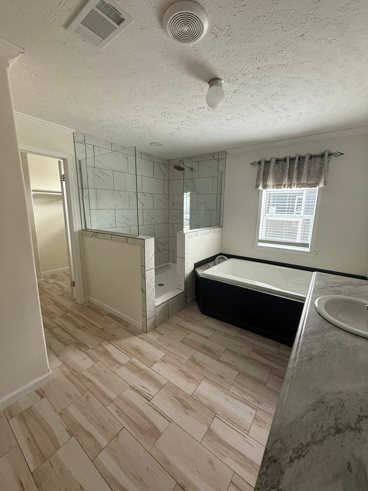 Bright bathroom featuring a marble-tiled shower with glass panels, a large black-framed bathtub, light wood flooring, and a window with curtains.
