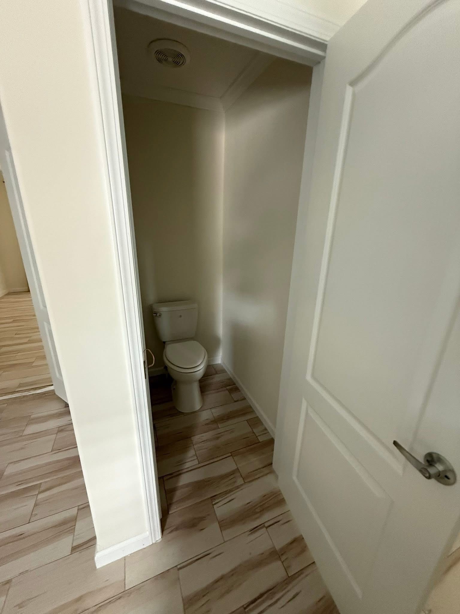 A small bathroom with a white toilet in a narrow space. The floor features light wooden tiles, and the door is partially open, creating a sense of simplicity.