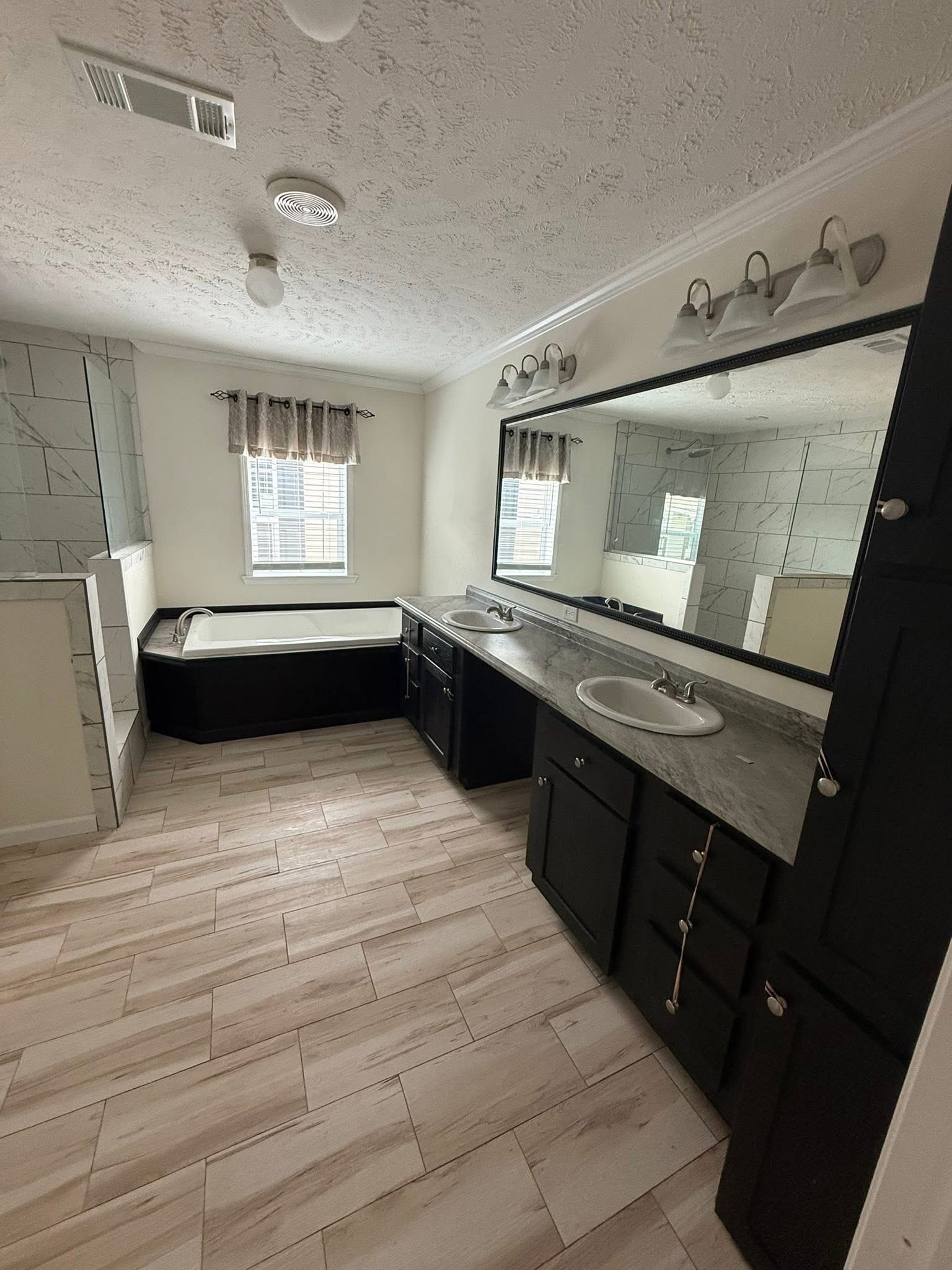 Spacious bathroom with dual sinks, large mirror, and dark cabinets. Features a white soaking tub by a window with light curtains. Neutral tone tiles.
