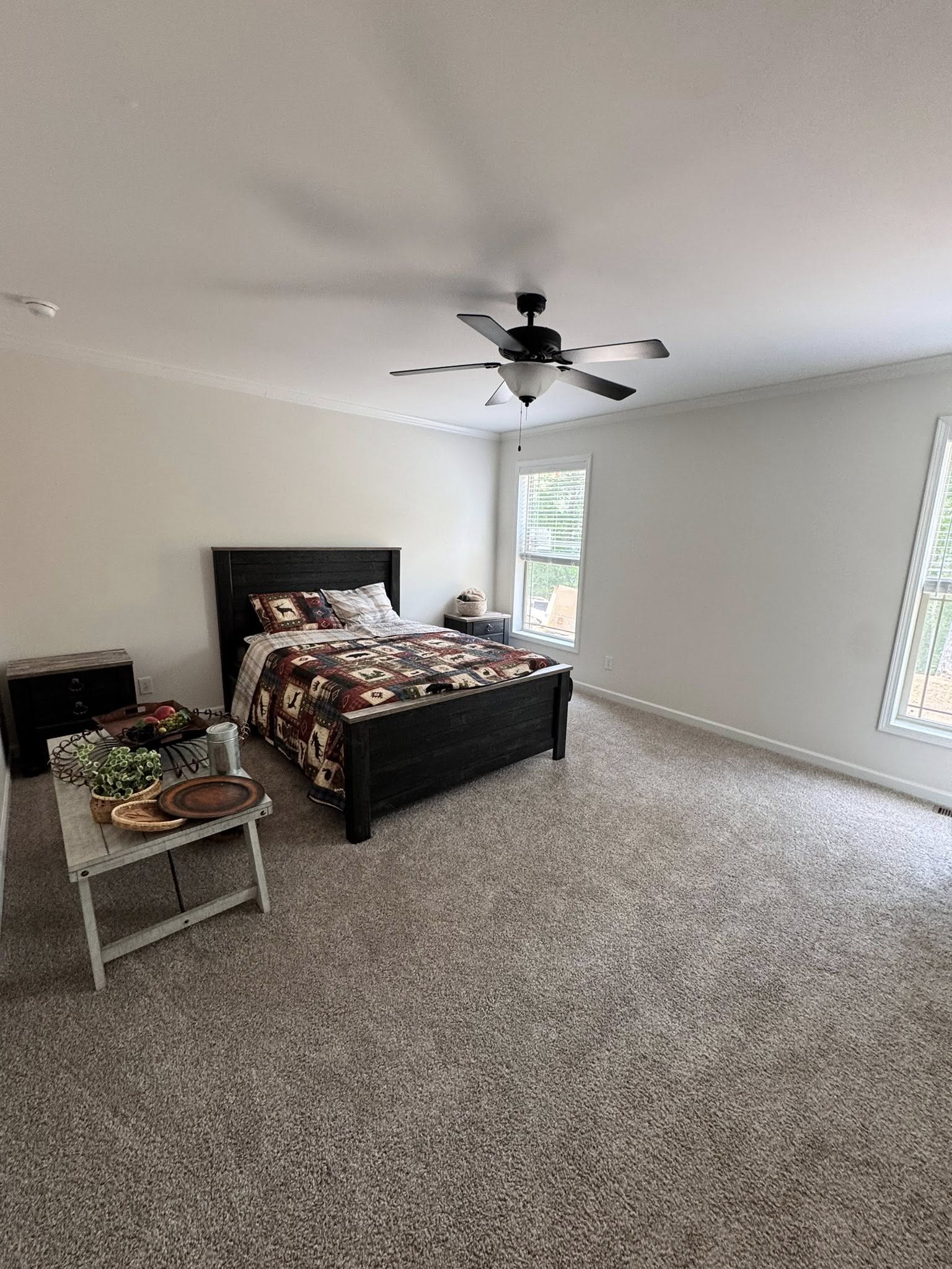 A cozy bedroom with a neatly made bed featuring a colorful patterned quilt. The room has white walls, two windows, and a gray carpet. A ceiling fan hangs above, and a wooden table with decorative items sits beside the bed, creating a warm and inviting atmosphere.