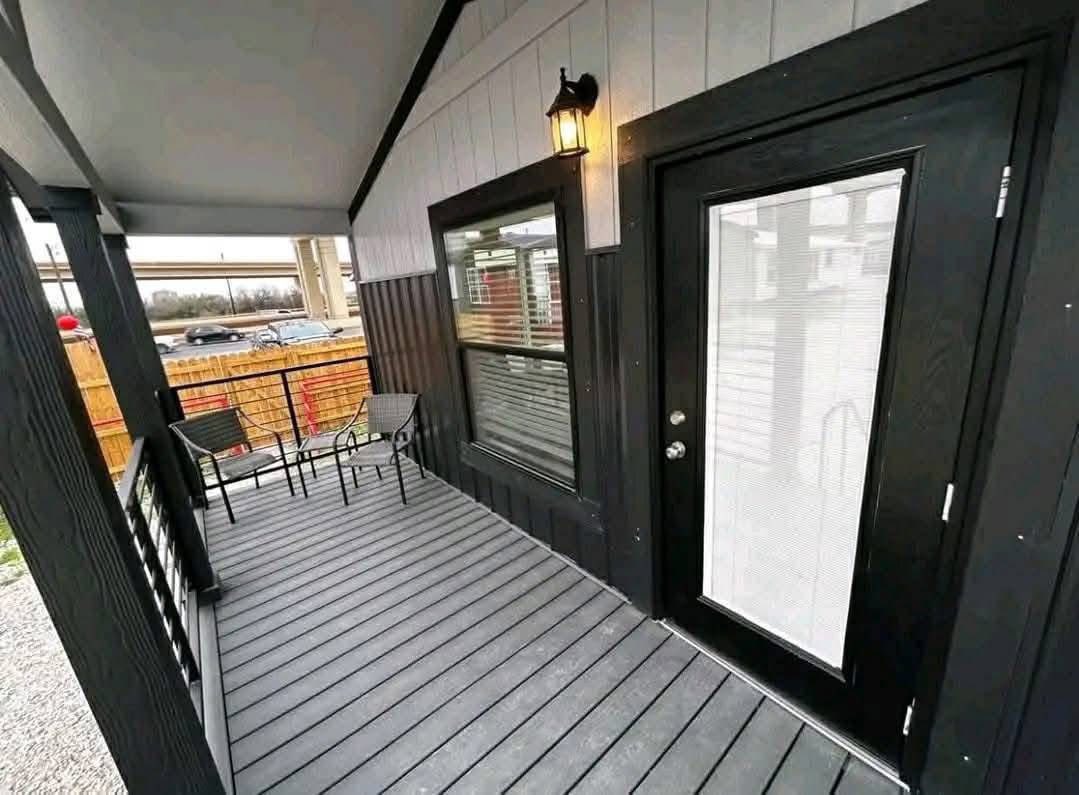 Covered porch with gray decking and black trim. Features black door, window, lamp, and patio furniture. Wooden fence and parked cars in background.
