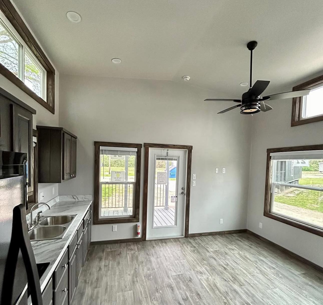 Bright kitchen with grey floors and dark cabinets, featuring a ceiling fan. Large windows and a glass door offer ample natural light, creating an airy feel.