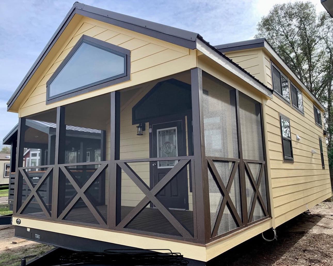 Yellow tiny house with brown trim, featuring a screened porch and triangular window. The cozy atmosphere is enhanced by surrounding greenery.