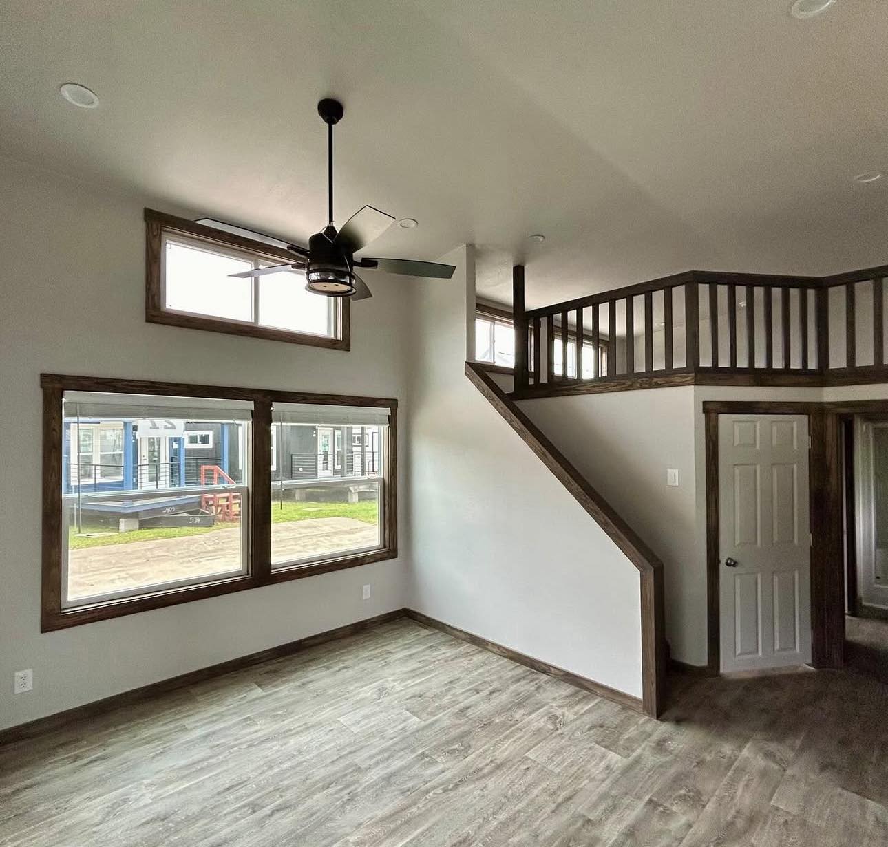 Modern minimalistic room with light gray walls and wooden flooring. Features large windows, a ceiling fan, and a loft with wooden railing.